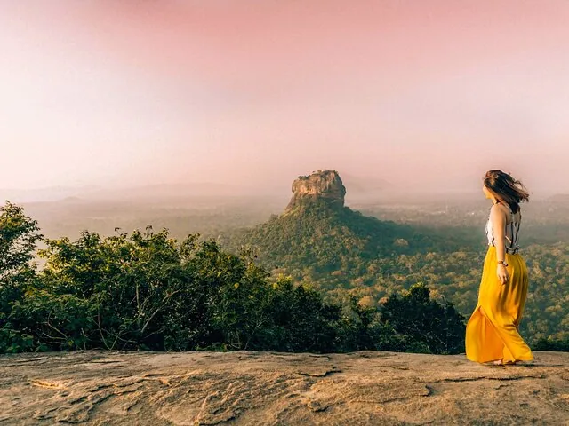 Frau in gelbem Rock blickt bei Sonnenaufgang vom Pidurangala-Felsen aus zum Sigiriya-Felsen