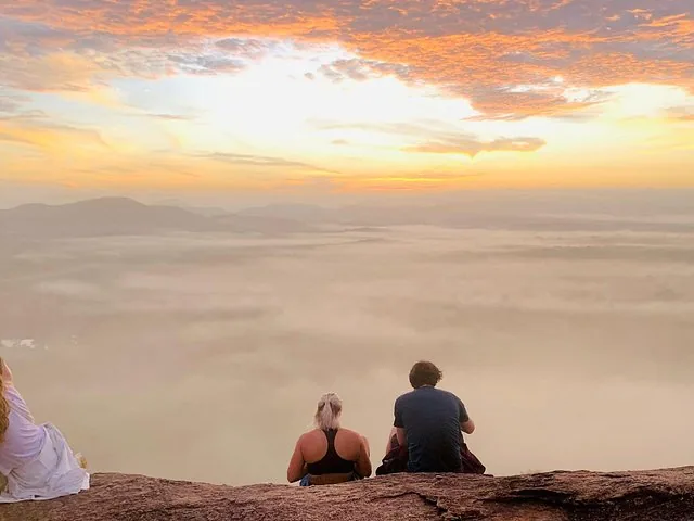 Menschen sitzen auf einer Felskante und beobachten eine neblige Landschaft im Sonnenaufgang